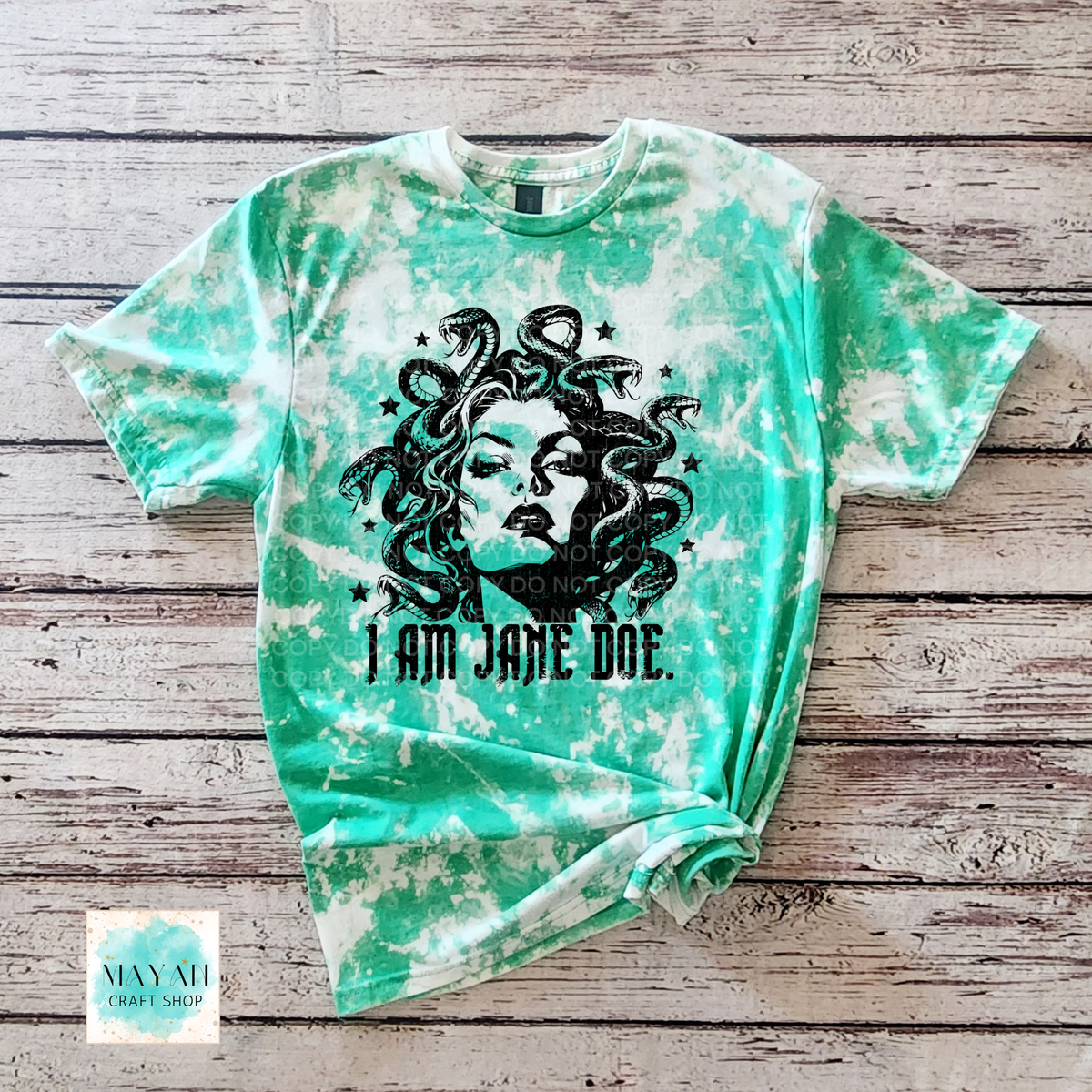 A heather irish green bleached t-shirt featuring a black graphic of Medusa with the text 'I AM JANE DOE.' printed below it, laid flat on a rustic wooden plank background.
