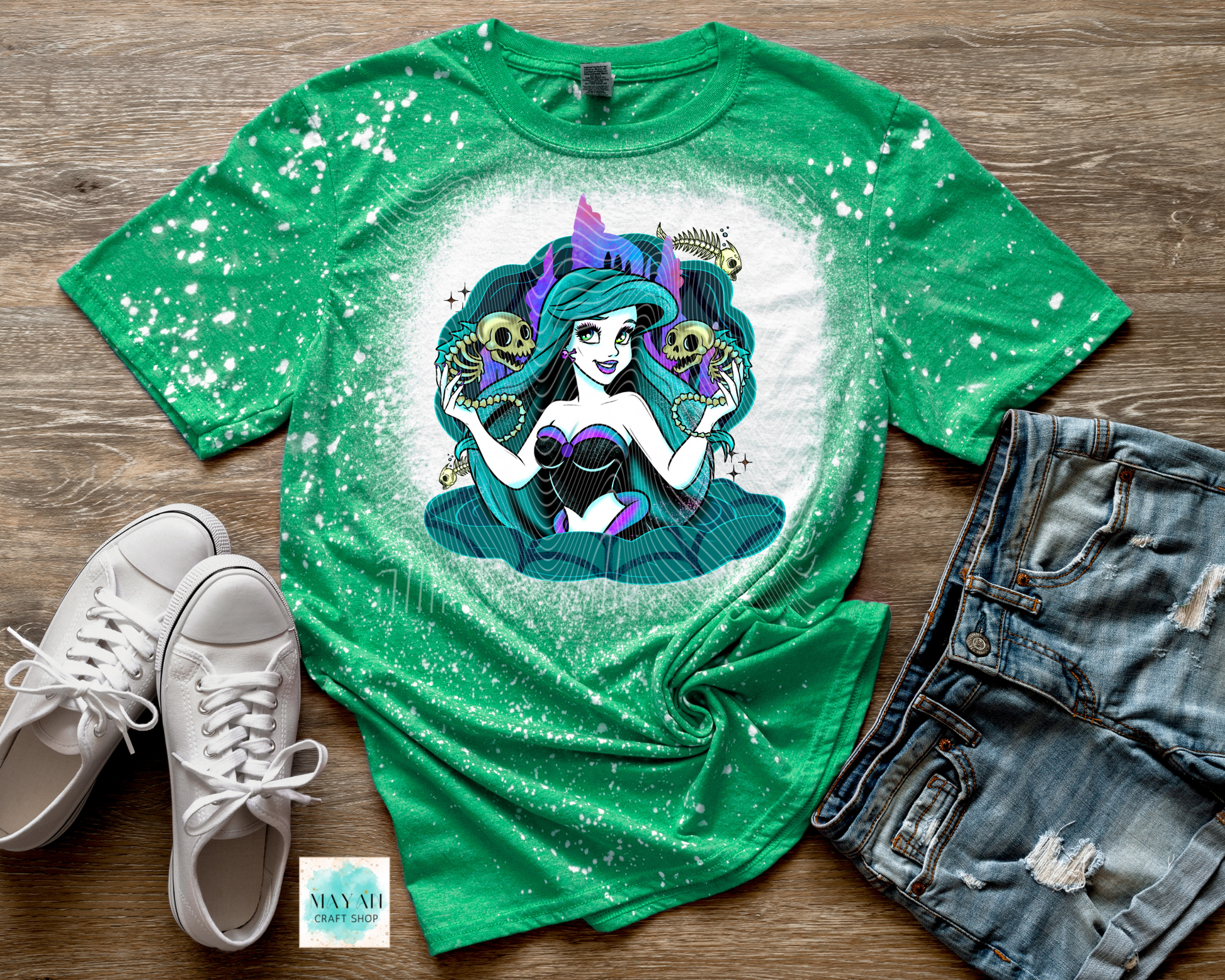 Green t-shirt with a mermaid graphic on a wooden surface with white sneakers and denim shorts.