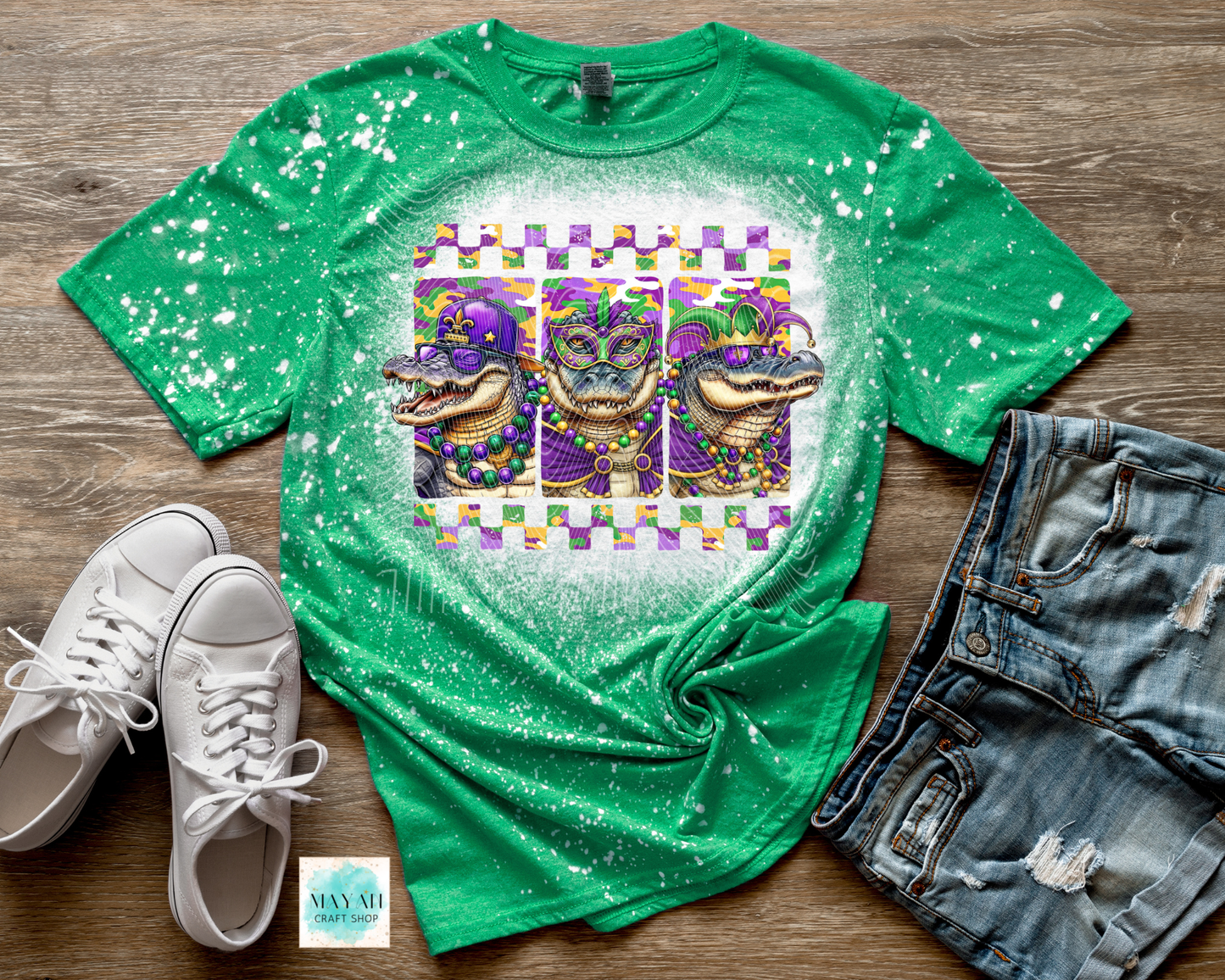 Green t-shirt with alligator Mardi Gras design on a wooden surface with white sneakers and denim shorts.