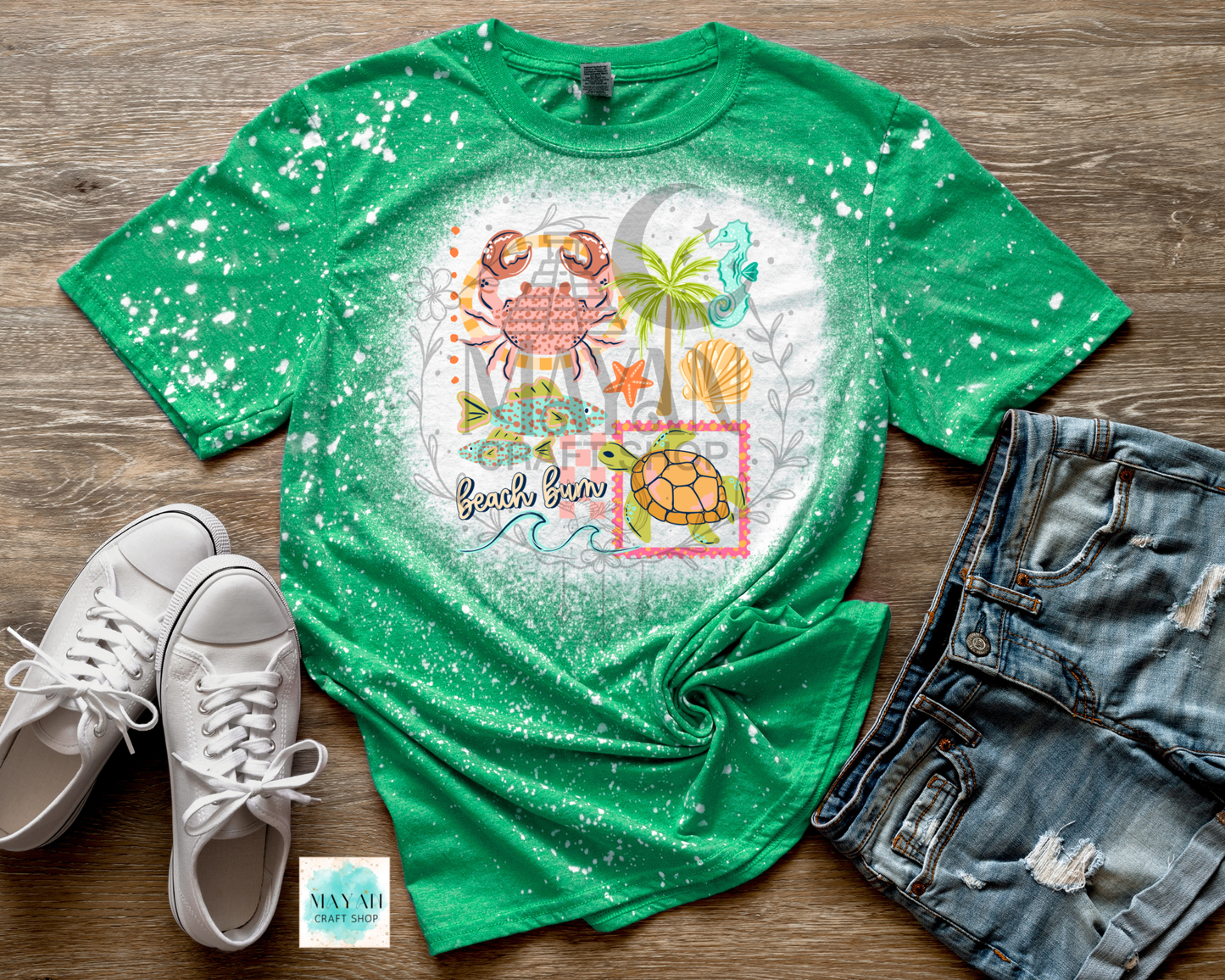 Green t-shirt with beach-themed design on a wooden surface with white sneakers and denim shorts.