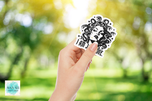 A hand holds a die-cut sticker featuring a black and white illustration of Medusa with the text 'I AM JANE DOE' on a blurred green outdoor background.