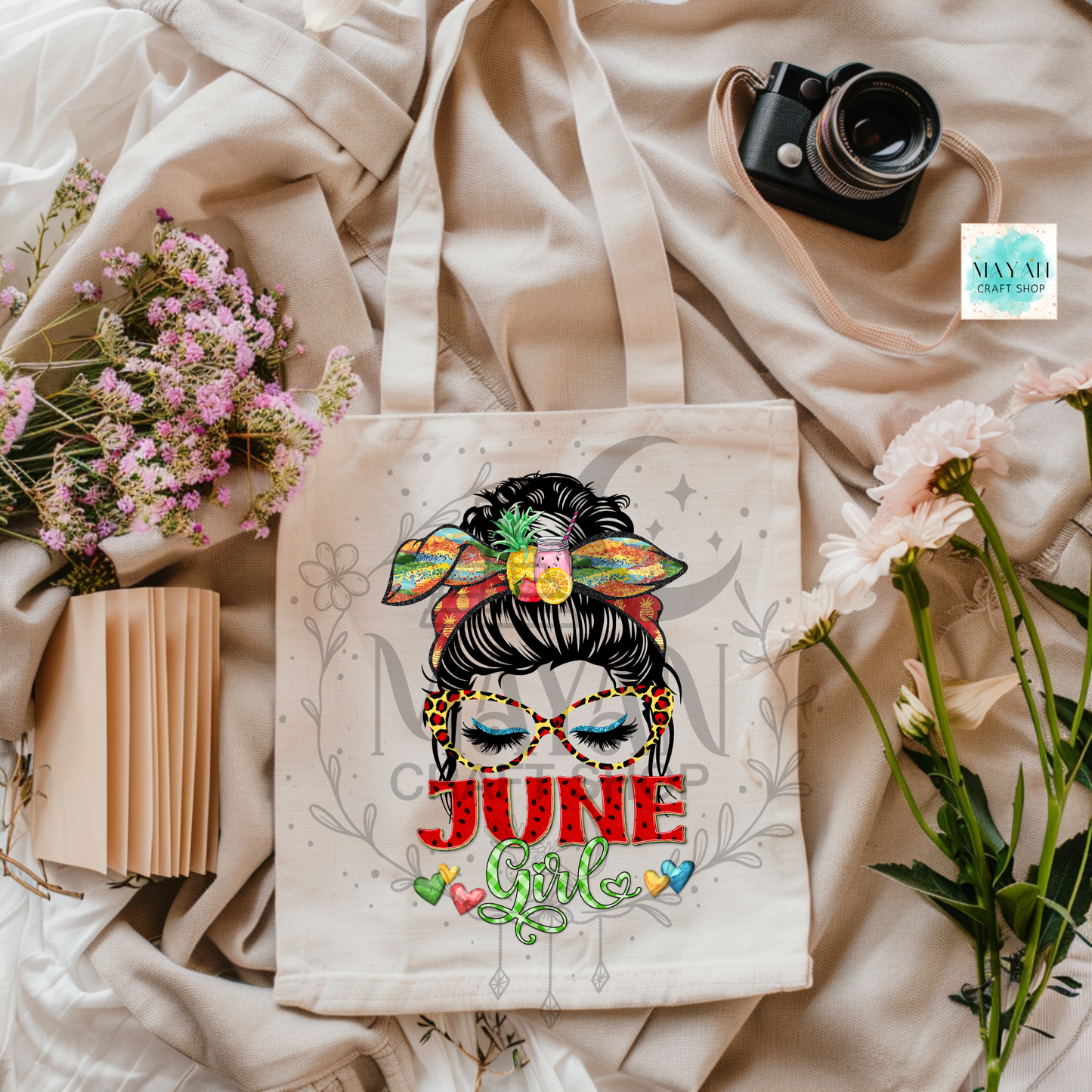 Tote bag with 'June Girl' design on a fabric background with flowers and a camera.