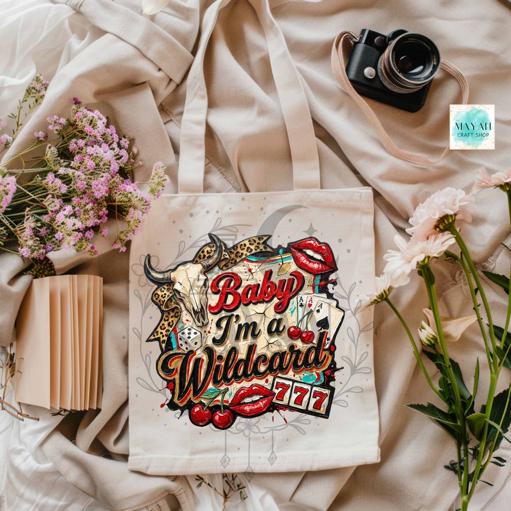 Tote bag with 'Baby I'm a Wildcard 777' design on a soft fabric background with flowers and a camera.