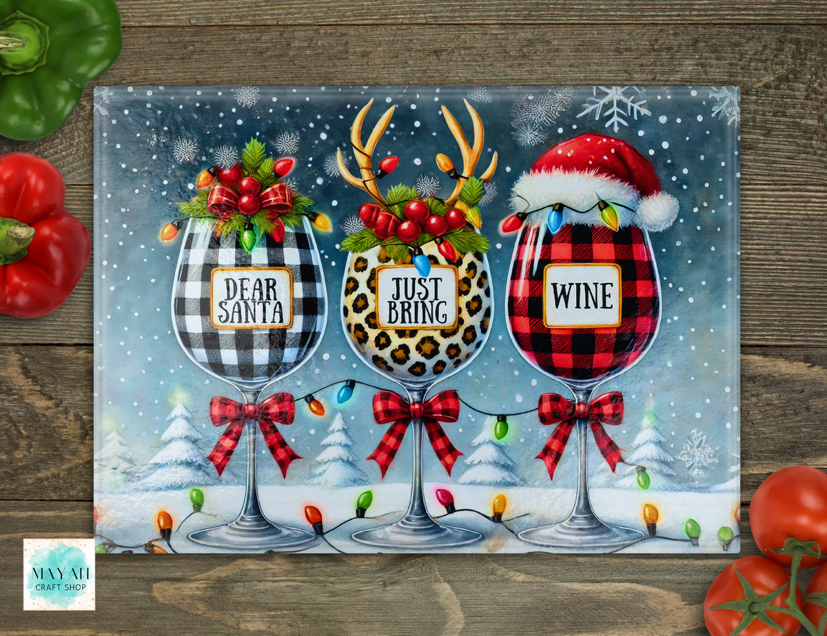 A cutting board featuring three wine glasses decorated for Christmas, with labels reading 'DEAR SANTA,' 'JUST BRING,' and 'WINE,' set against a snowy winter scene. The sign is photographed flat on a rustic wood surface with red and green peppers around it.