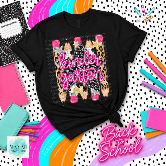 A black graphic t-shirt featuring a 'Kinder Garten' school supply design with pencils in leopard print and pink erasers, styled in a flat-lay with school supplies and colorful curved stripes.