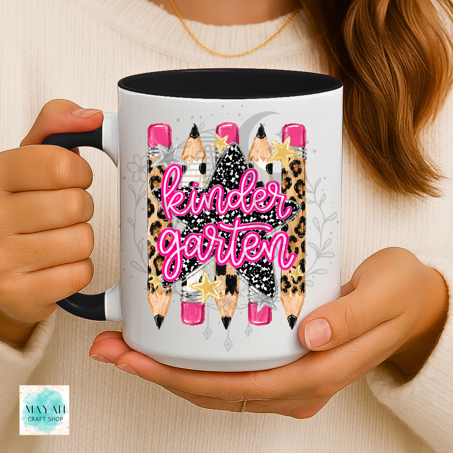 A person holds a white ceramic mug with a black handle and interior. The mug features a graphic of colorful pencils with leopard-print and black-and-white patterns, stars, and the words 'kindergarten' in bright pink script.
