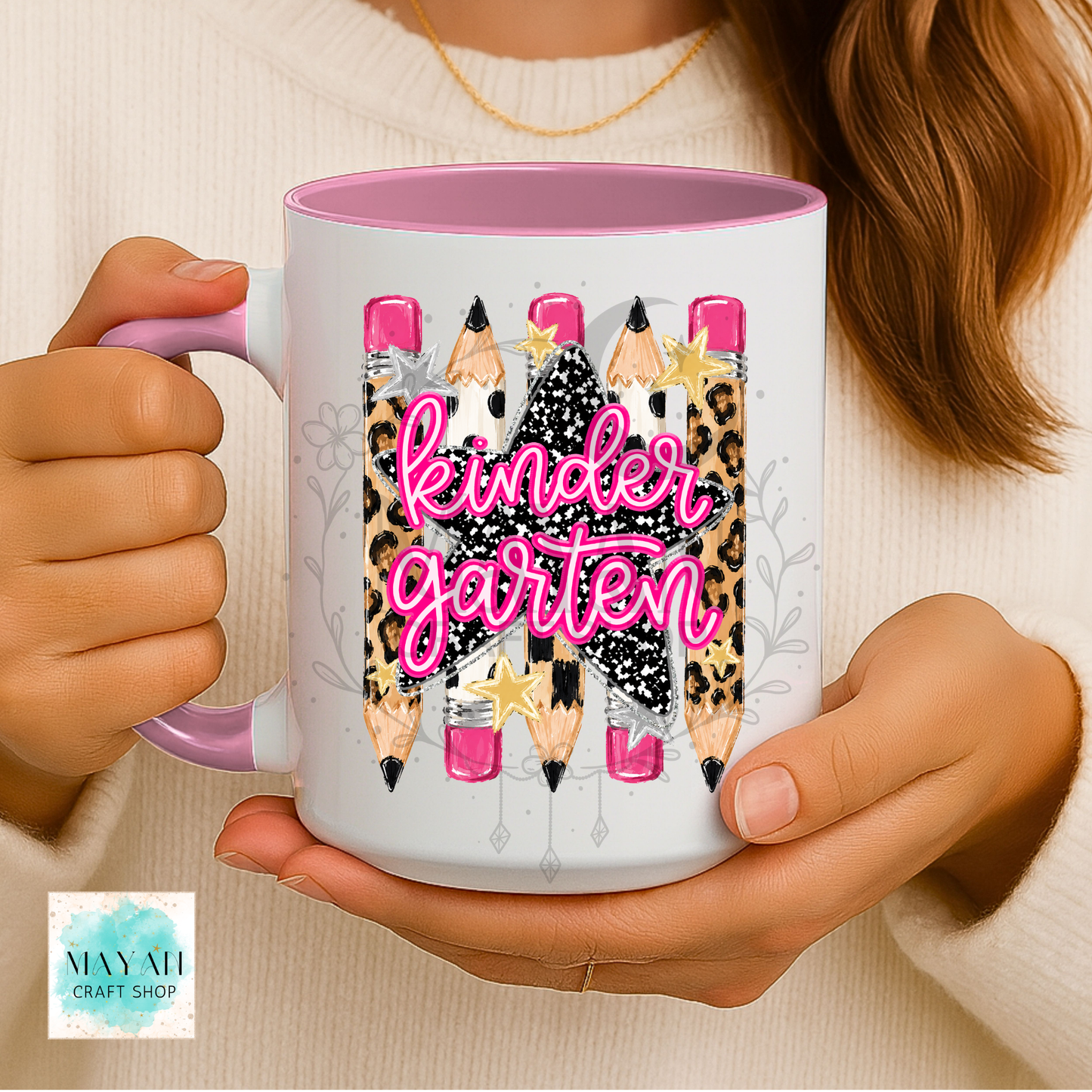 A person holds a white ceramic mug with a pink interior and handle. The mug features a graphic of five pencils with leopard print, black-and-white patterned skirts, pink erasers, and the words 'kindergarten' in pink script.