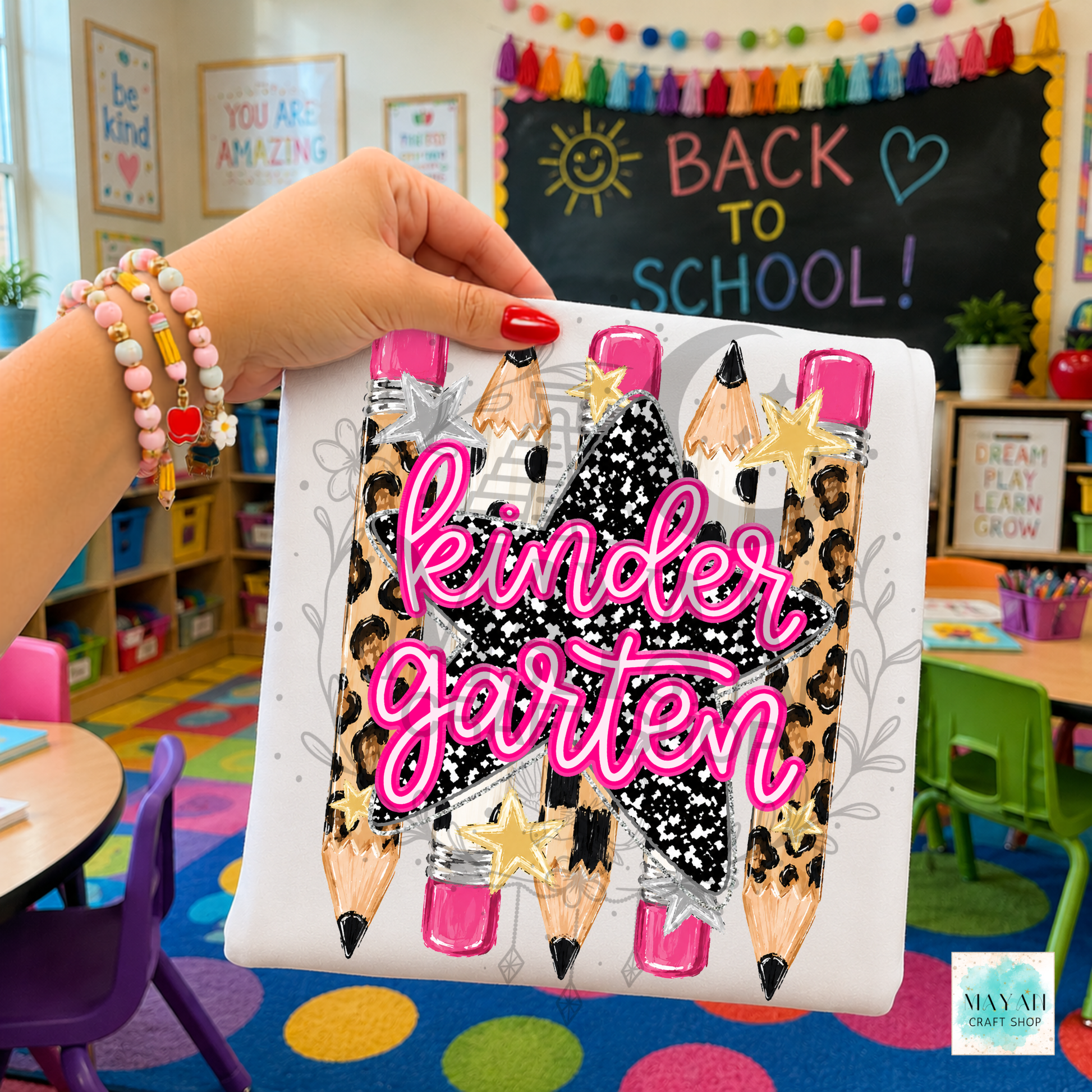 A hand holds a white shirt featuring a design of four pencils with leopard print, black glitter patterns, pink and gold erasers, and the text 'kinder garten' in bright pink script. The background shows a colorful children's classroom with a chalkboard that says 'BACK TO SCHOOL!'.