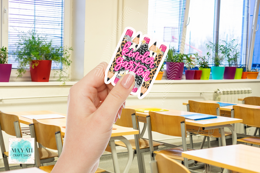 A hand holds a die-cut sticker featuring three pink and black leopard-print pencils, set against a blurred classroom background with desks and potted plants.