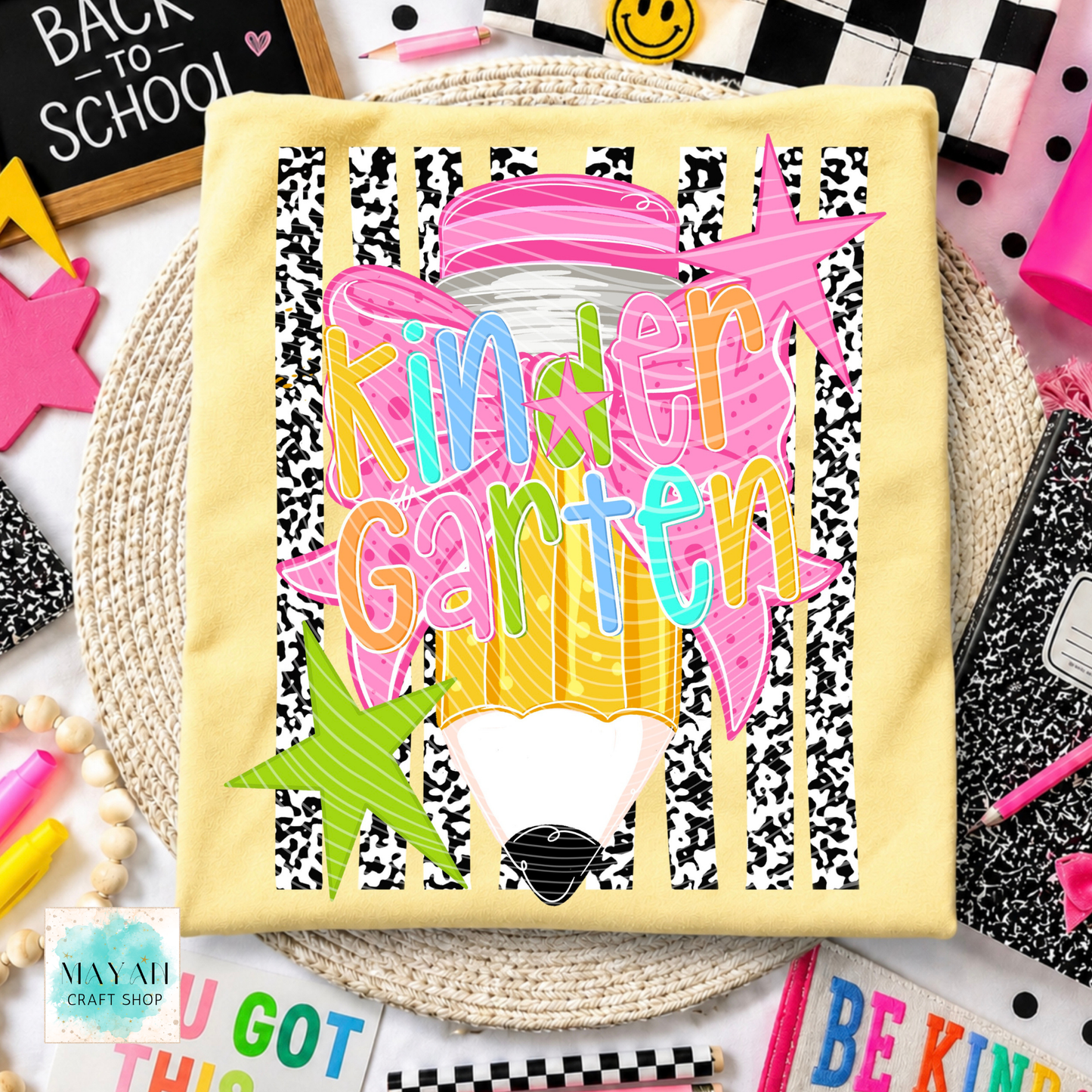 A square pale yellow shirt, likely a napkin or small cloth square, featuring a colorful 'Kinder Garten' school-themed graphic with a pencil, stars, and black-and-white stripes, styled on a woven placemat with school supplies.