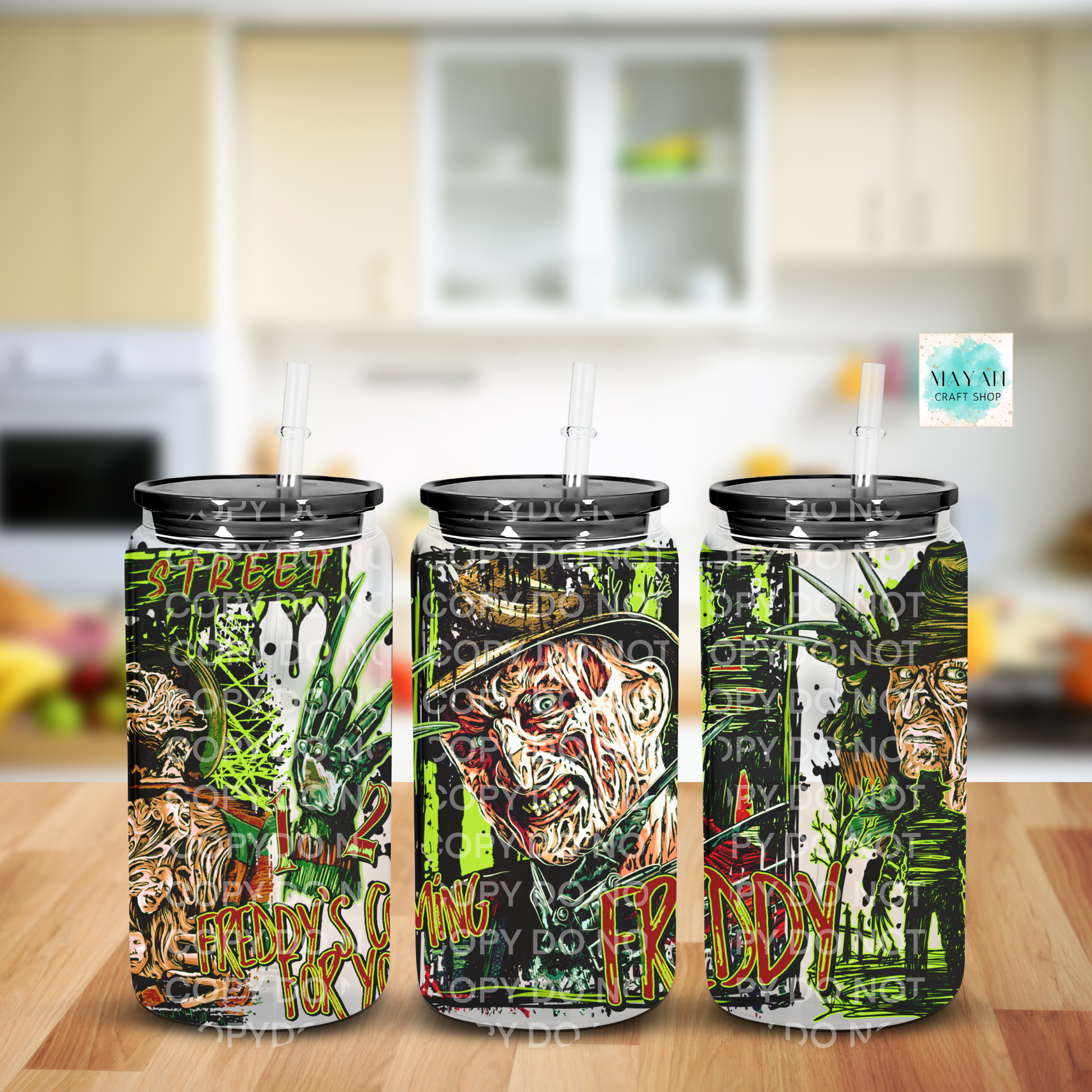 Three tall skinny tumblers with black lids and clear straws, printed with a horror-themed collage and green graffiti-style text, displayed on a wooden surface with a blurred kitchen background.