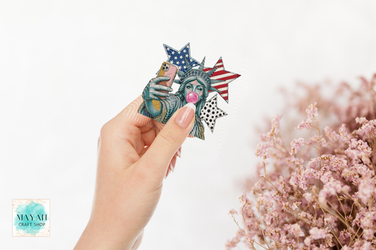Hand holding a decorative item with a design of the Statue of Liberty and American flag, with pink flowers in the background.