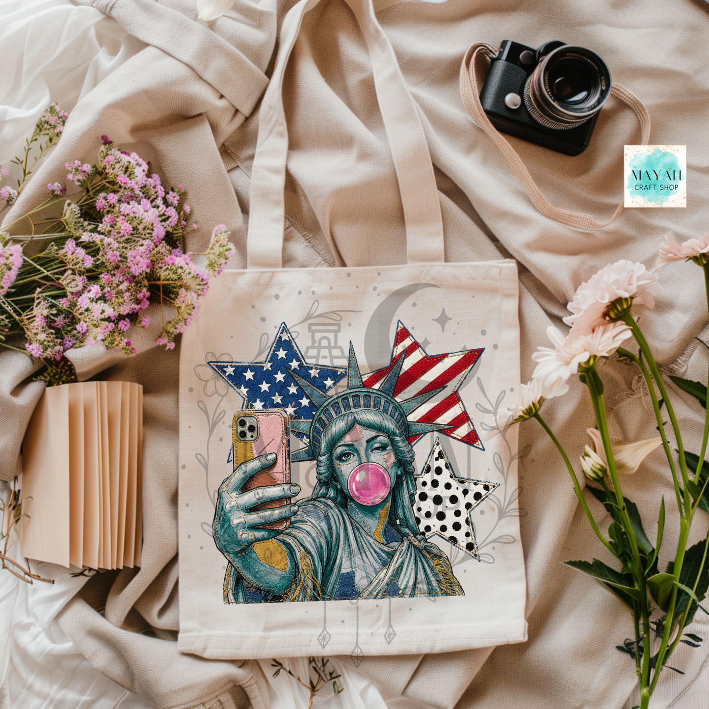 Tote bag with a stylized Statue of Liberty holding a phone, surrounded by flowers and a camera on a textured surface.