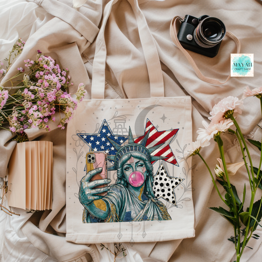 Tote bag with a stylized Statue of Liberty holding a phone, surrounded by flowers and a camera on a textured surface.