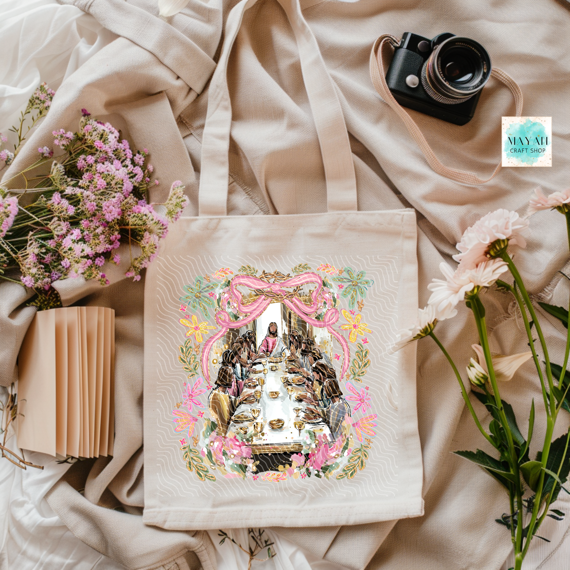Tote bag with artistic design on a soft fabric background with flowers and a camera.