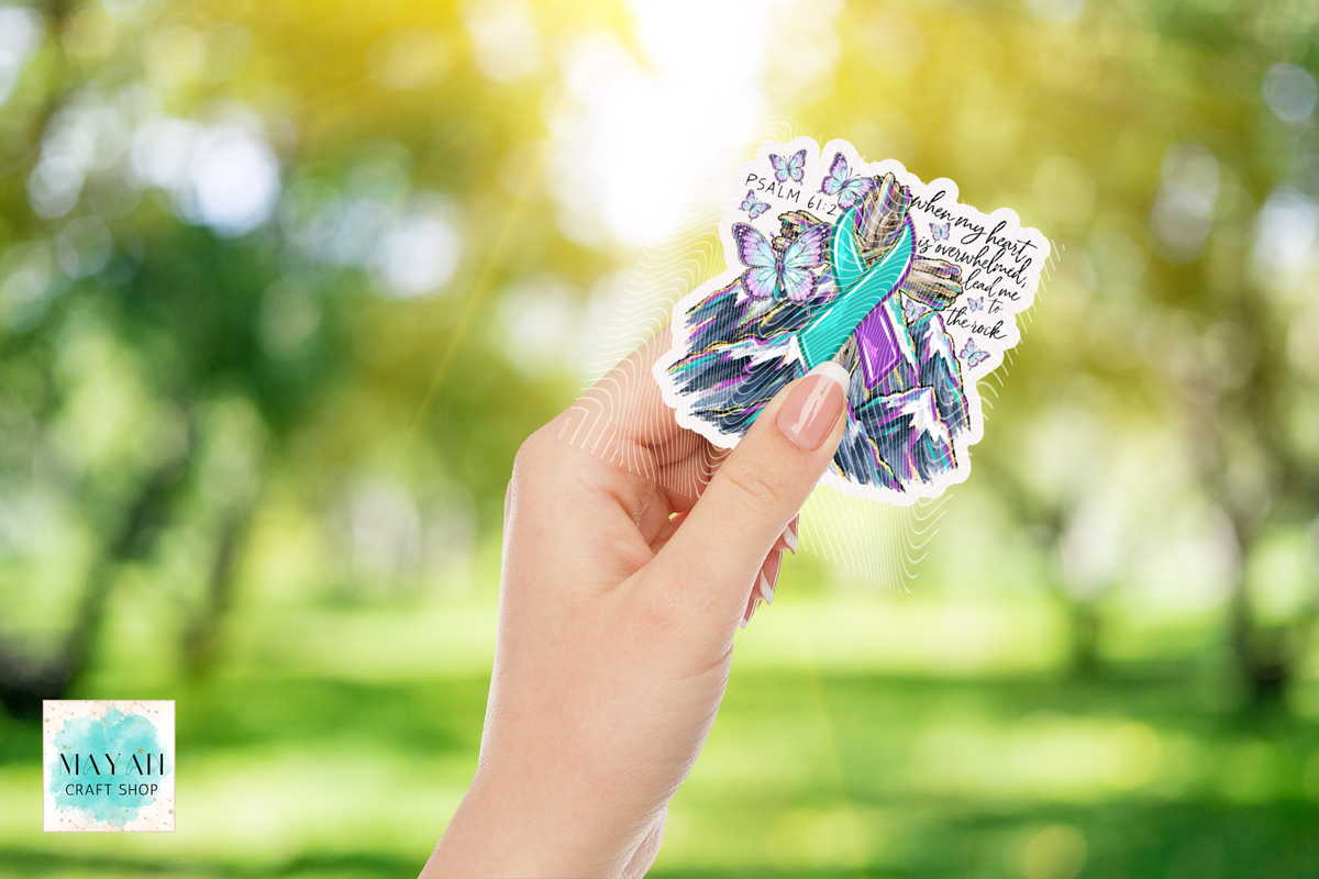 A hand holds a die-cut sticker featuring a colorful ribbon design and inspirational text, set against a blurred green outdoor background. 