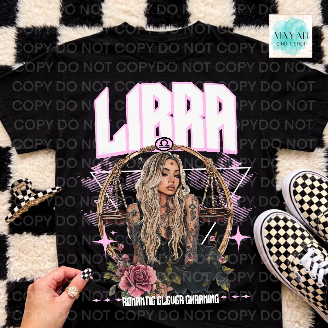 A black graphic T-shirt laid flat on a black-and-white checkered rug, featuring a Libra zodiac illustration with the word 'LIBRA' in large pink letters, a woman's portrait, scales, roses, and decorative text. A hand is visible at the lower left, and black-and-white checkered sneakers appear at the lower right.