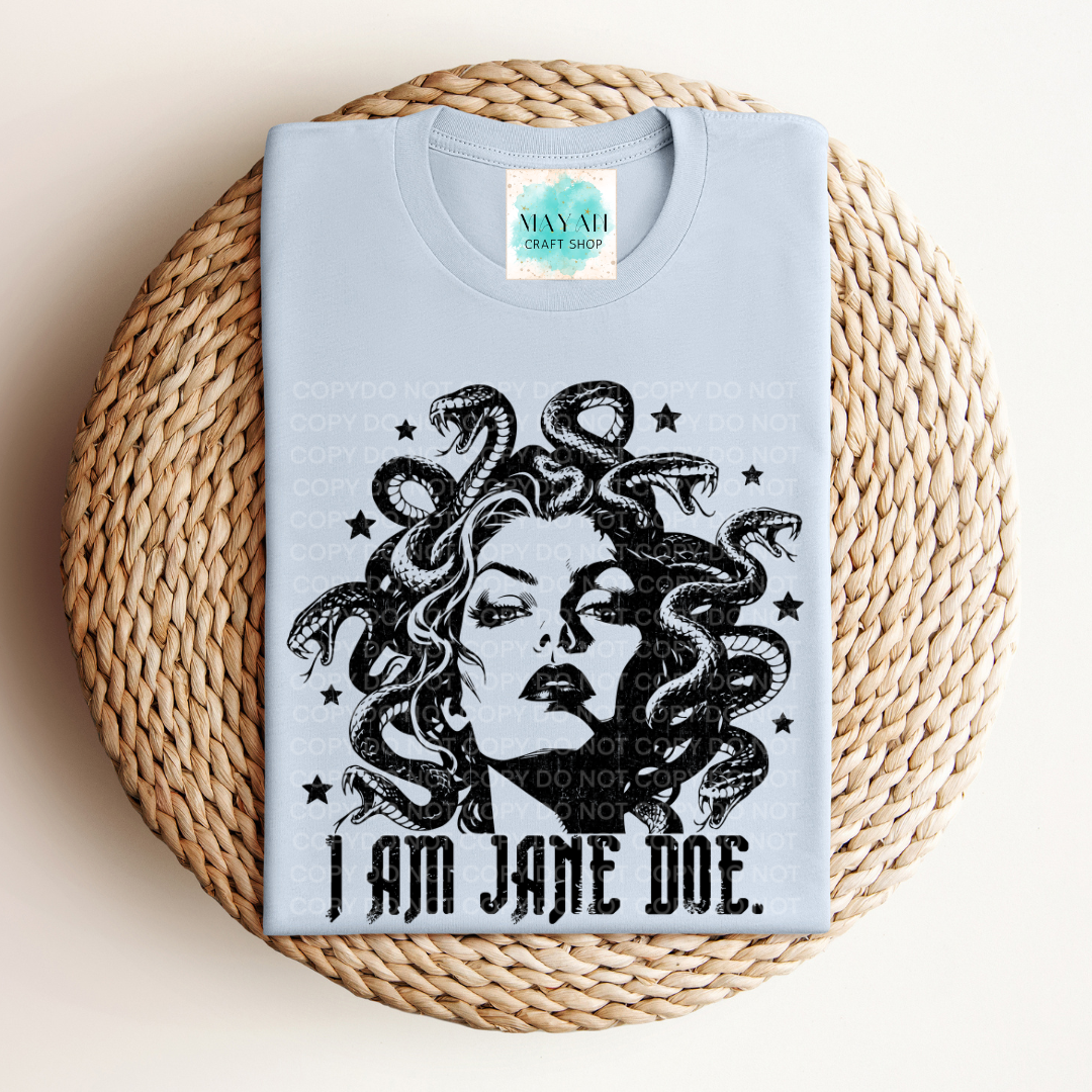 A folded light blue graphic T-shirt displayed on a round woven straw mat. The shirt features a black illustration of Medusa with snakes in her hair and the text 'I AM JANE DOE.