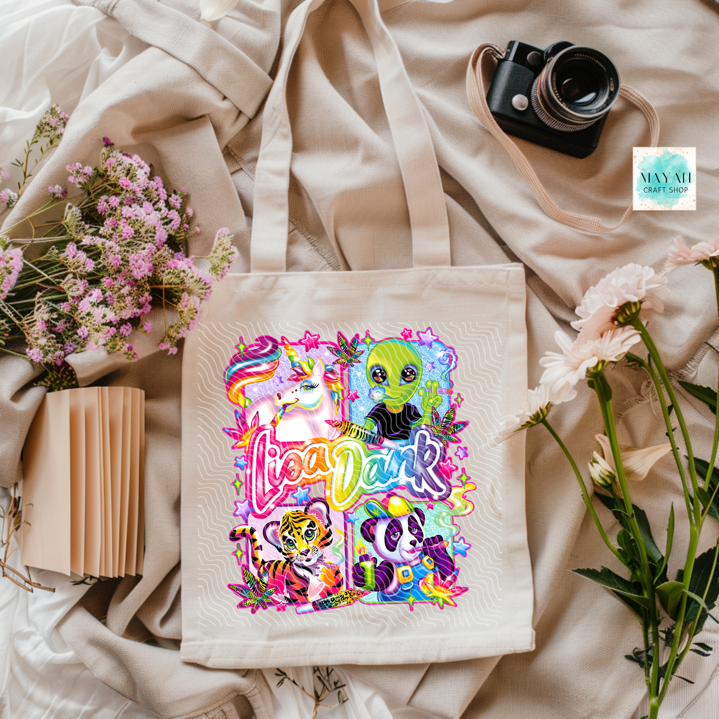 Tote bag with colorful design featuring characters and text, placed on a light fabric background with flowers and a camera.
