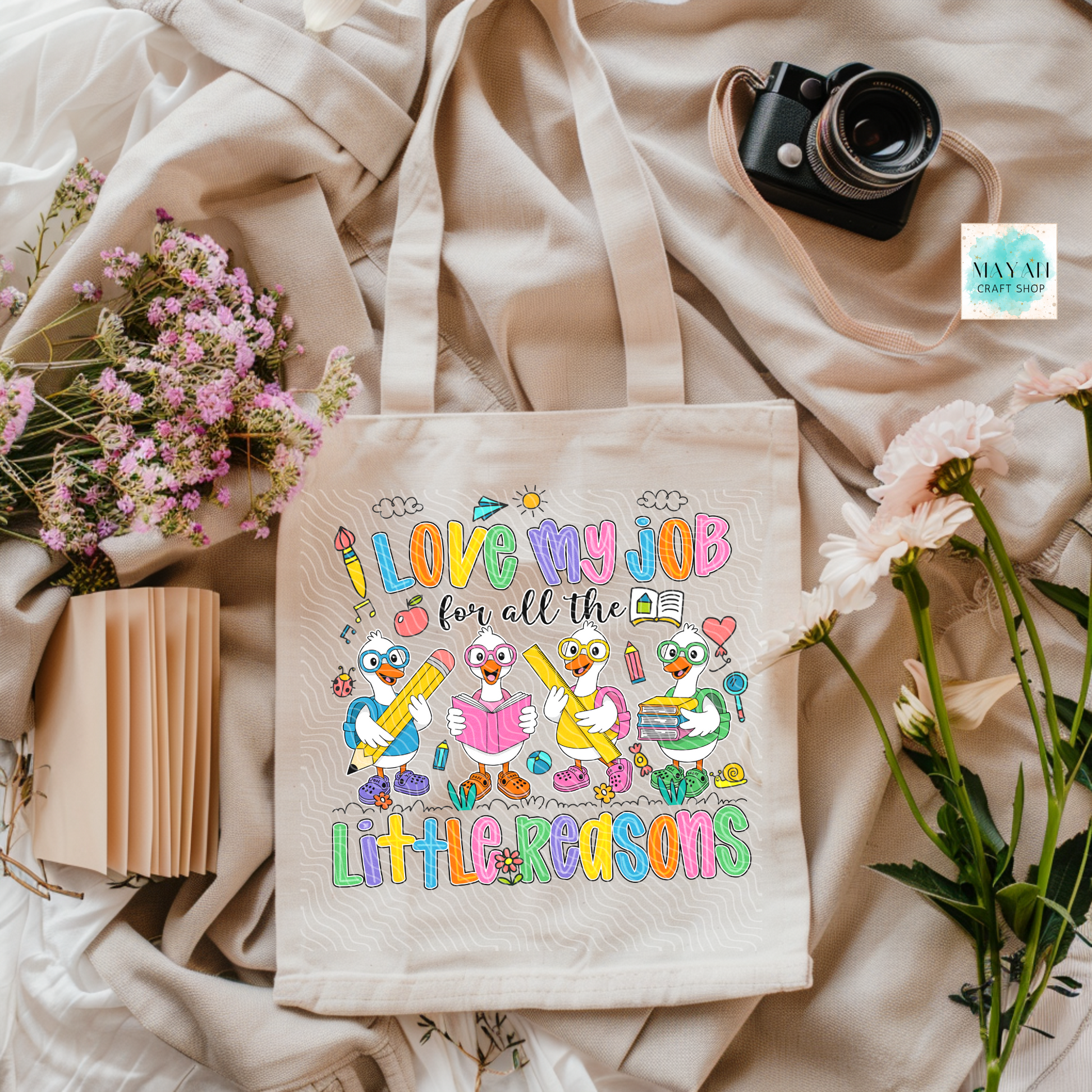 Tote bag with colorful design and text on a soft fabric background with flowers and a camera.