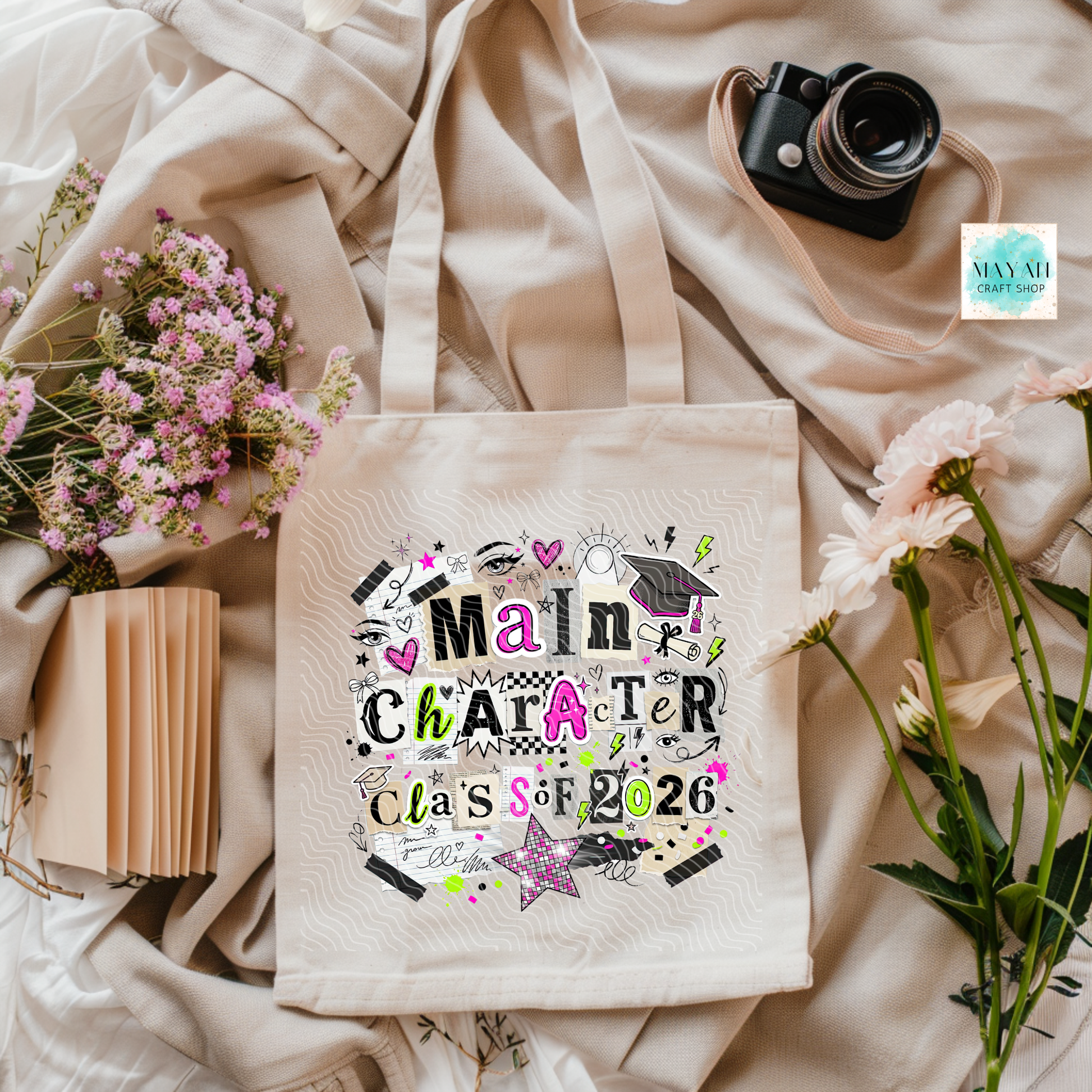 Tote bag with 'Main Character Class of 2026' design on a soft fabric background with flowers and a camera.
