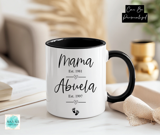 White mug with black interior and handle displaying 'Mama Est. 1981 Abuela Est. 1997' text on a blurred home background.