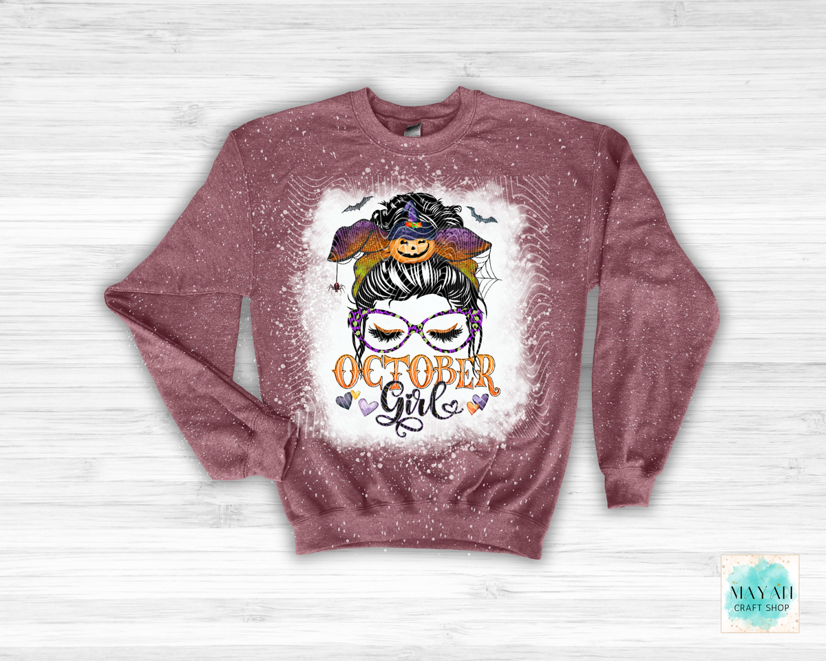 A dusty rose crewneck sweatshirt with a distressed, glittery texture, featuring a central graphic of a witch with a pumpkin hat and the text 'OCTOBER Girl' on a white splattered background, displayed flat on a white wood plank surface.