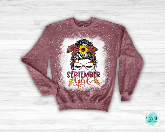 A heather maroon crewneck sweatshirt with a 'September Girl' graphic, laid flat on a white wood background. The graphic features a woman with glasses, a sunflower in her hair, and autumn leaves, with the text 'SEPTEMBER Girl' in pink and orange.