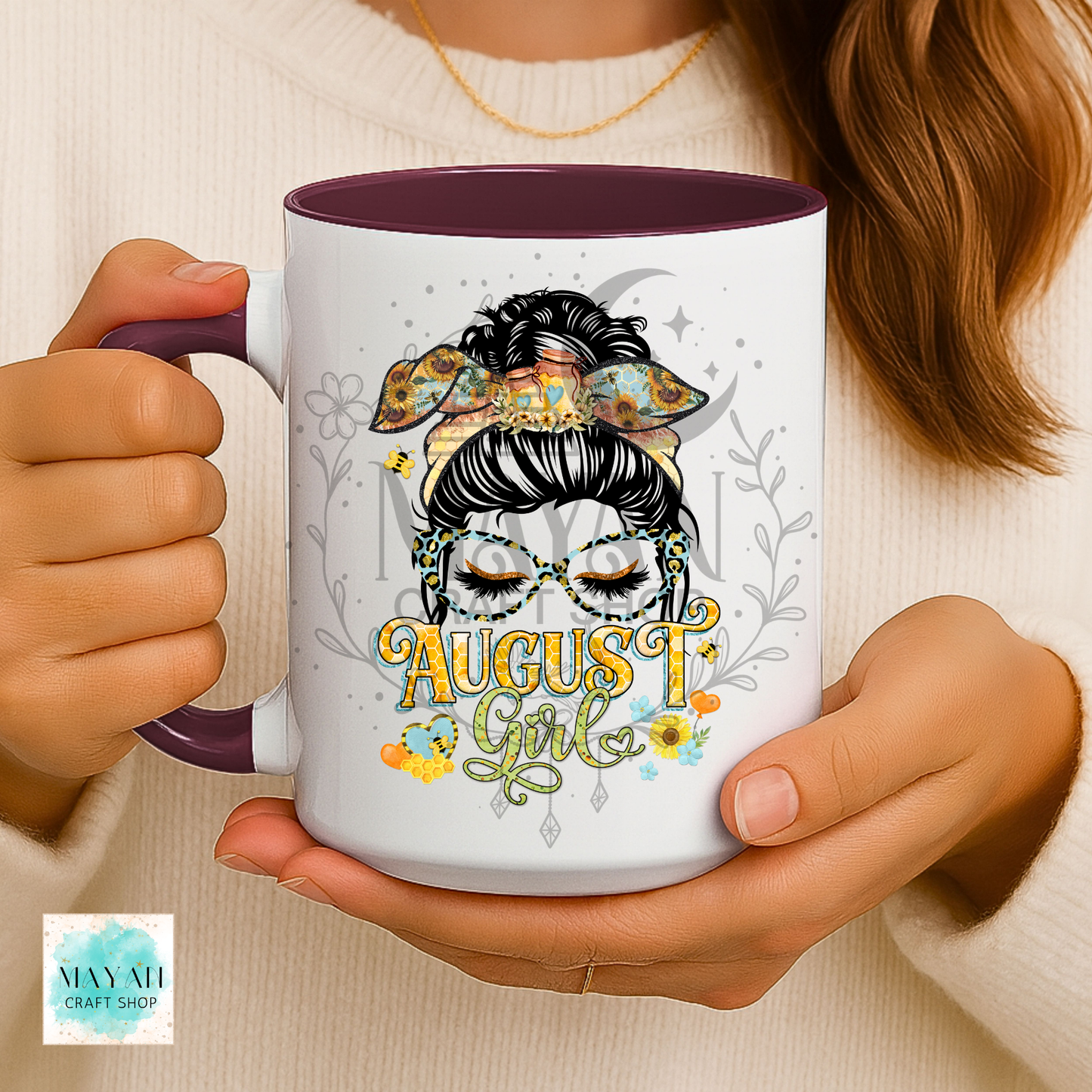 A person holds a white ceramic mug with a dark purple interior and handle. The mug features a graphic of a woman with glasses and a bow, with the text 'AUGUST Girl' in yellow and green script, surrounded by small floral details.
