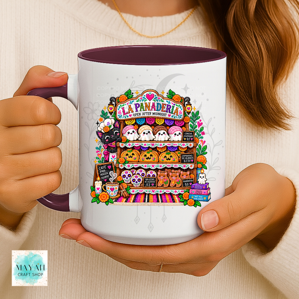 A person holds a white ceramic mug with a maroon handle and interior. The mug features a colorful illustration of a Halloween-themed bakery display with jack-o'-lanterns, ghosts, pumpkins, and the text 'LA PANADERIA'.