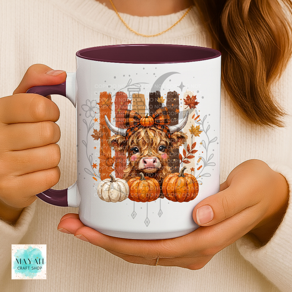A person holds a white ceramic mug with a dark purple interior and handle. The mug features a fall-themed illustration of a Scottish Highland cow, pumpkins, autumn leaves, and a fence.