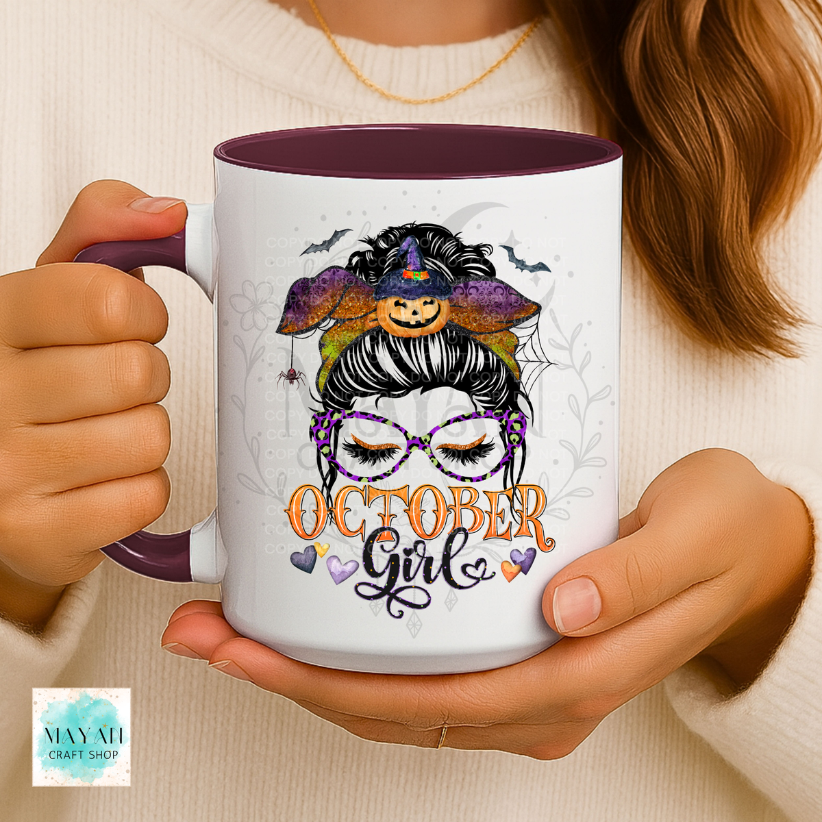 A person holds a white ceramic mug with a dark purple interior and handle. The mug features a Halloween-themed graphic of a woman with glasses, a witch hat, and the text 'October Girl' in orange and purple.