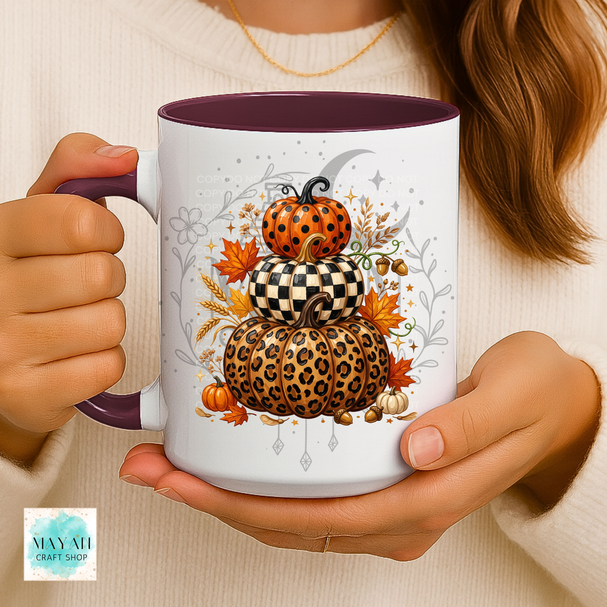 A person holds a white ceramic mug with a dark purple handle and interior. The mug features a stacked autumn pumpkin illustration with leopard print, checkerboard pattern, and orange pumpkins, surrounded by fall leaves and acorns.