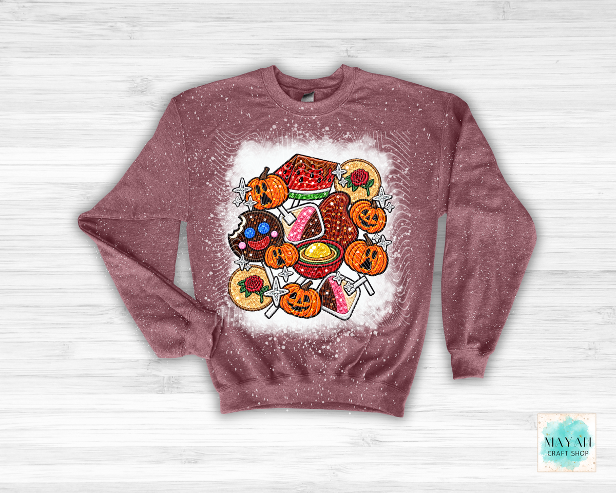 A heather maroon long-sleeve sweatshirt with a white distressed graphic print of various Halloween treats, including jack-o'-lanterns, candy corn, a watermelon watermelon, and a chocolate chip cookie, laid flat on a white wood background.