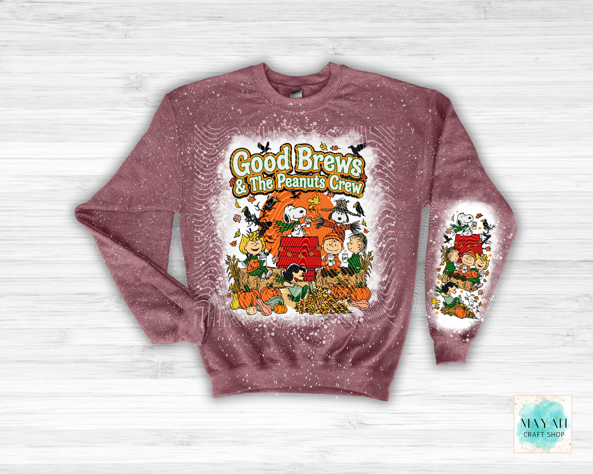 A long-sleeve crewneck sweatshirt in a heather maroon, featuring a festive 'Good Brews' graphic on the front and a matching graphic on the left sleeve. The sweatshirt is displayed flat against a light gray wood-plank background.