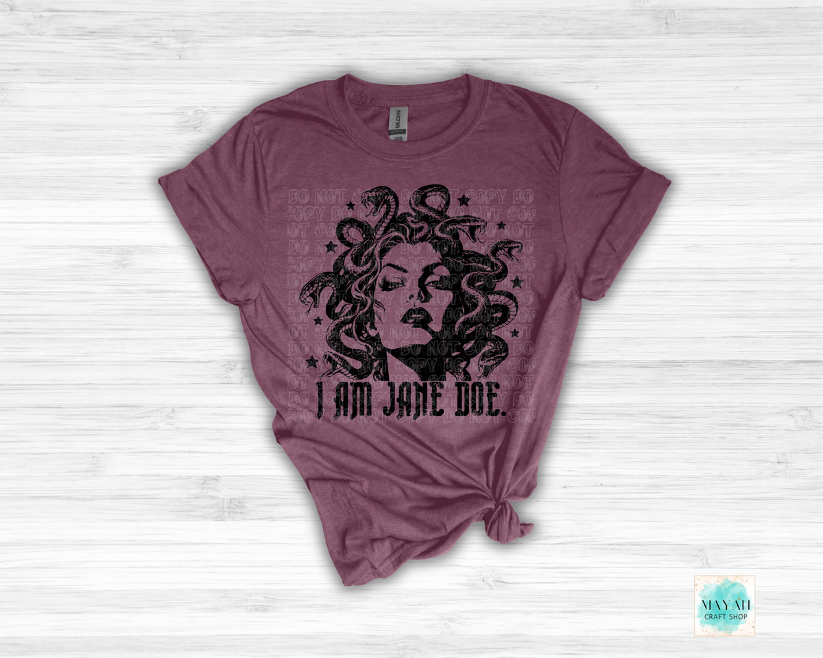 A heathered maroon short-sleeve t-shirt with a black graphic of Medusa and the text 'I AM JANE DOE.' printed on the front, styled flat on a white wood background.