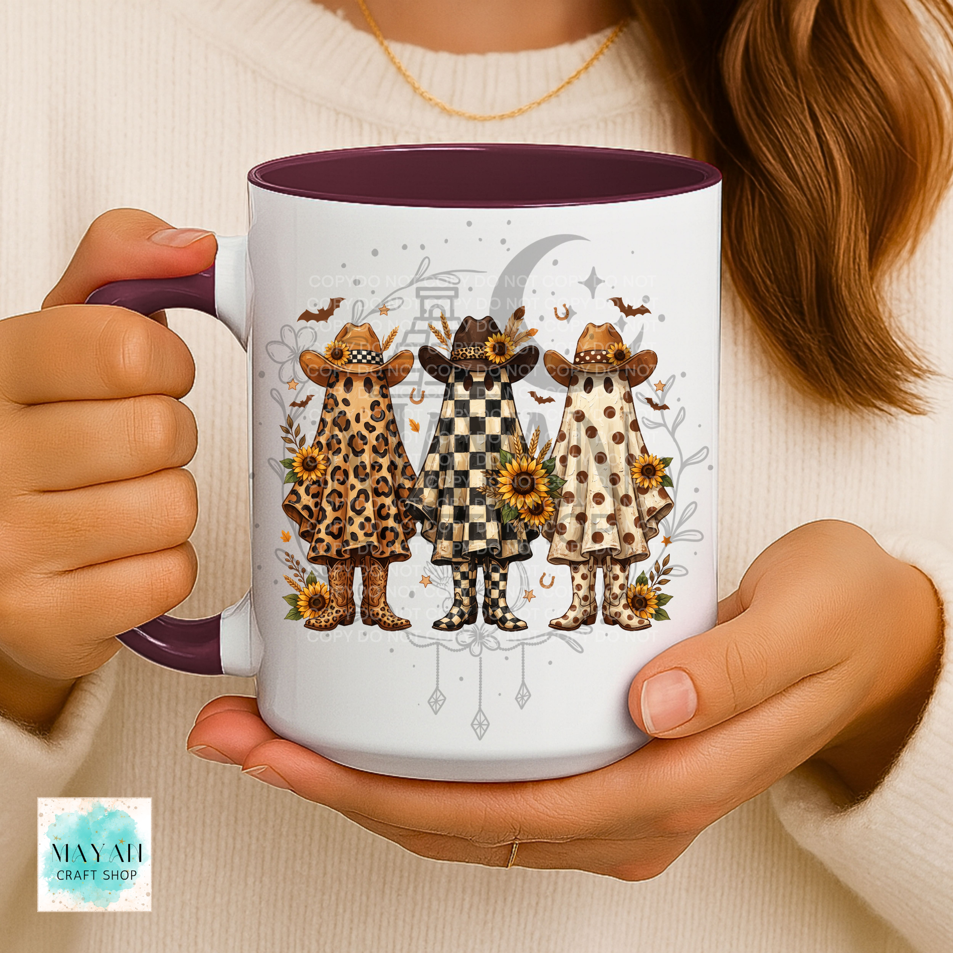 A person holds a white ceramic mug with a dark purple interior and handle. The mug features a printed illustration of three whimsical characters in animal-print cloaks and hats under a crescent moon and sunflowers.