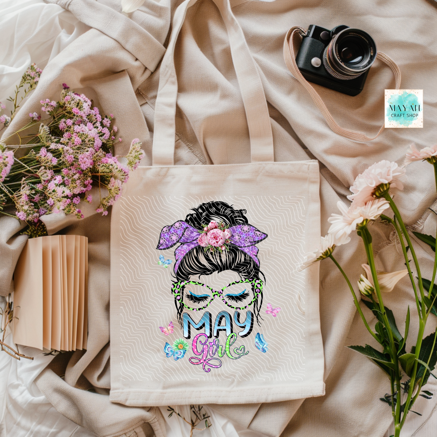 Tote bag with 'May Girl' design on a soft fabric background with flowers and a camera.