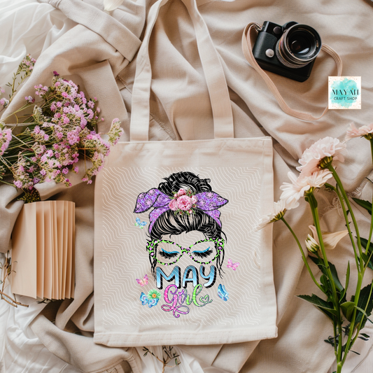 Tote bag with 'May Girl' design on a soft fabric background with flowers and a camera.