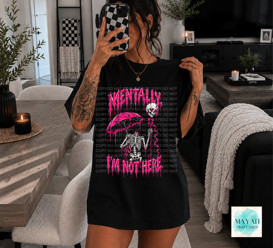 A woman takes a mirror selfie in a modern living room while wearing an oversized black graphic T-shirt. The shirt features a pink dripping design with the words 'MENTALLY I'M NOT HERE' and a skeleton holding a pink umbrella.