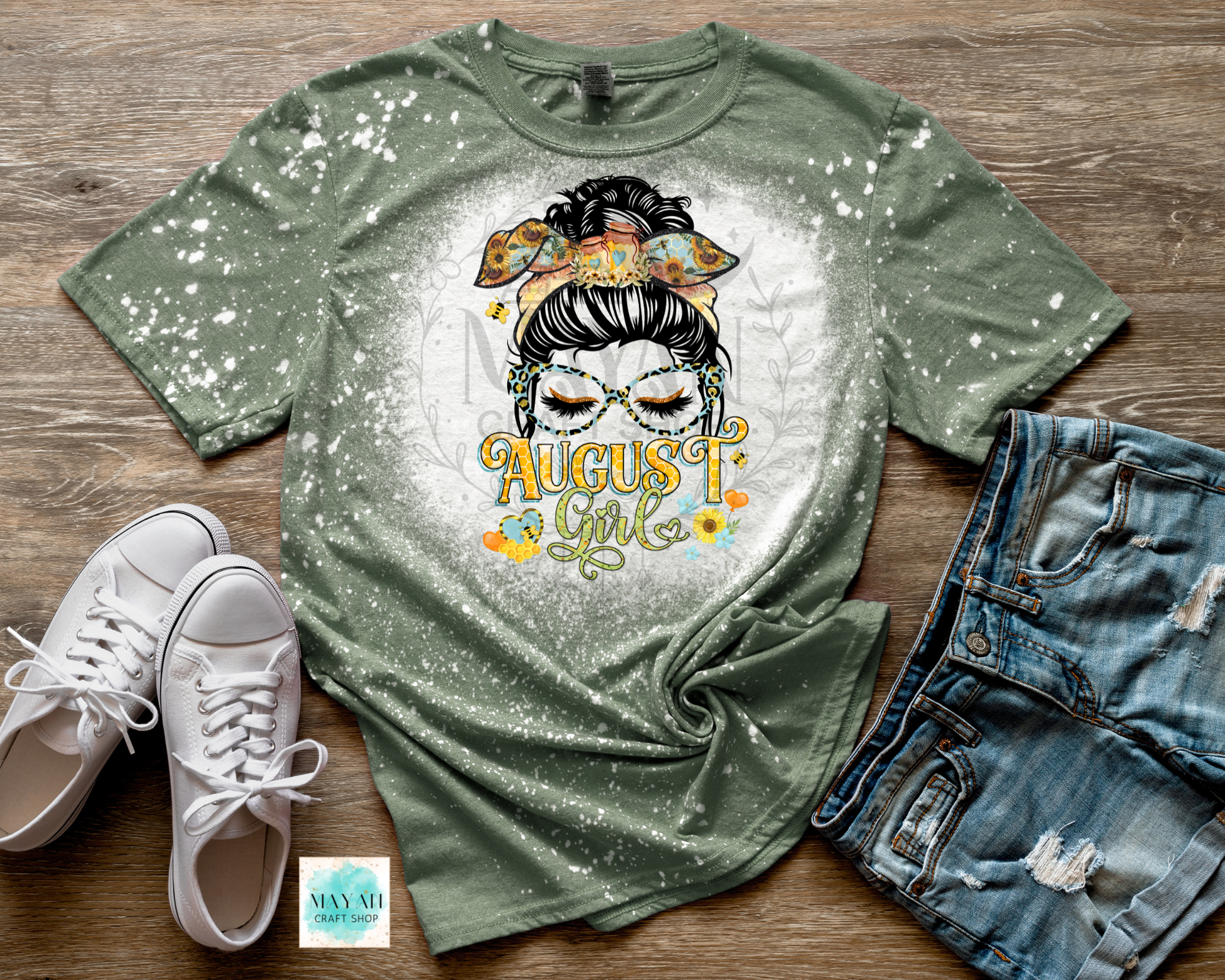 An olive green short-sleeve t-shirt with a white bleach-splatter finish and a graphic of a woman with glasses and the text 'August Girl' in yellow and orange. The shirt is styled in a flat lay on a wooden surface alongside white sneakers, distressed denim shorts.