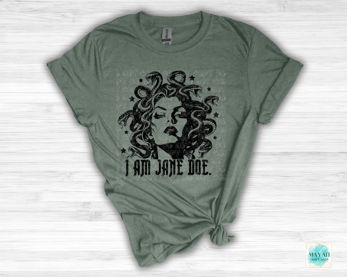 An heather military green short-sleeve t-shirt with a distressed black graphic of Medusa and the text 'I AM JANE DOE.' printed on the front, styled on a white wood background.