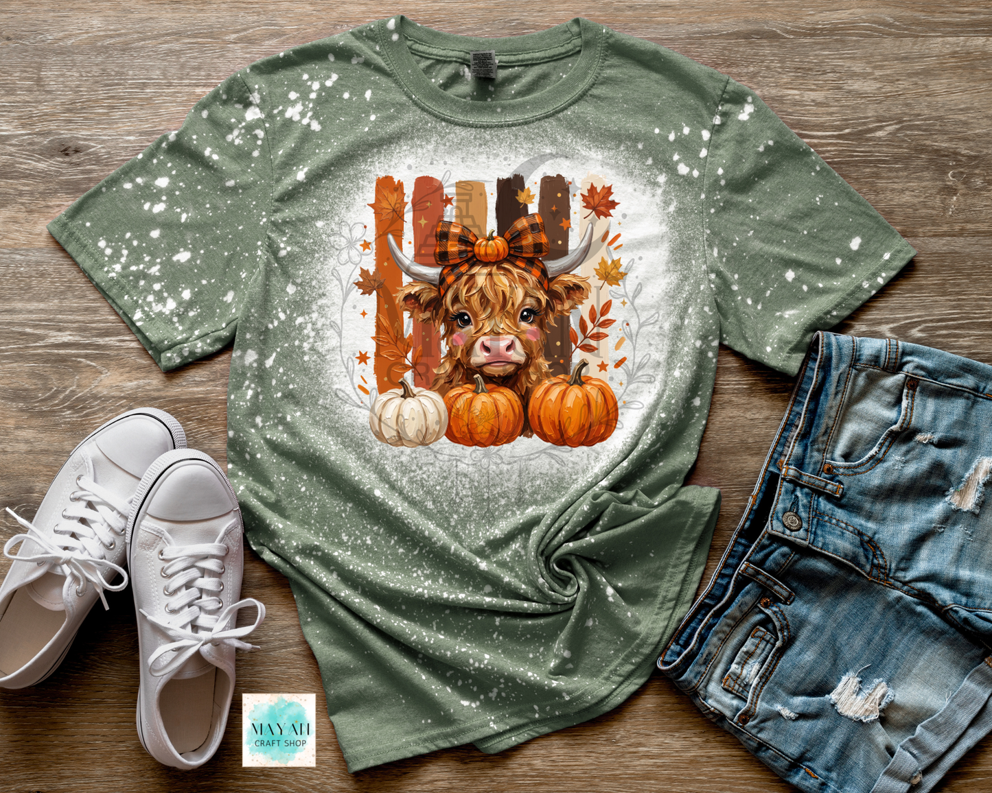 A heather military green t-shirt with a white speckled distressed finish, featuring a graphic of a Scottish highland cow in front of a wooden fence with pumpkins and autumn leaves. The shirt is styled in a flat lay on a wooden background with white sneakers, denim shorts.