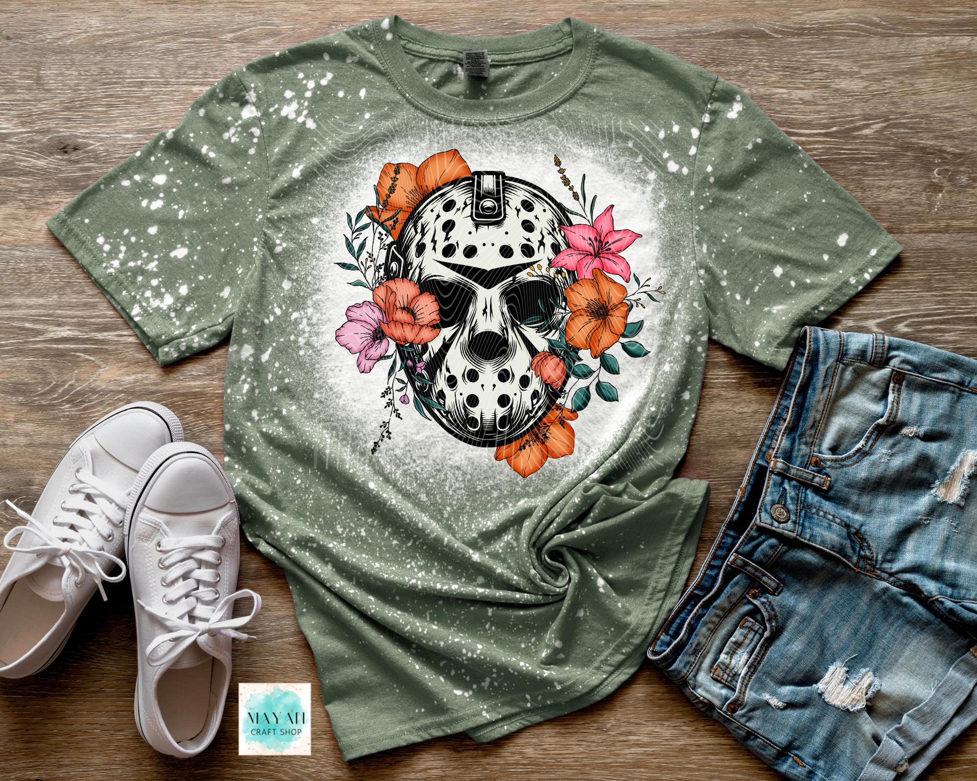 Green t-shirt with skull and floral design on a wooden surface with white sneakers and denim shorts.