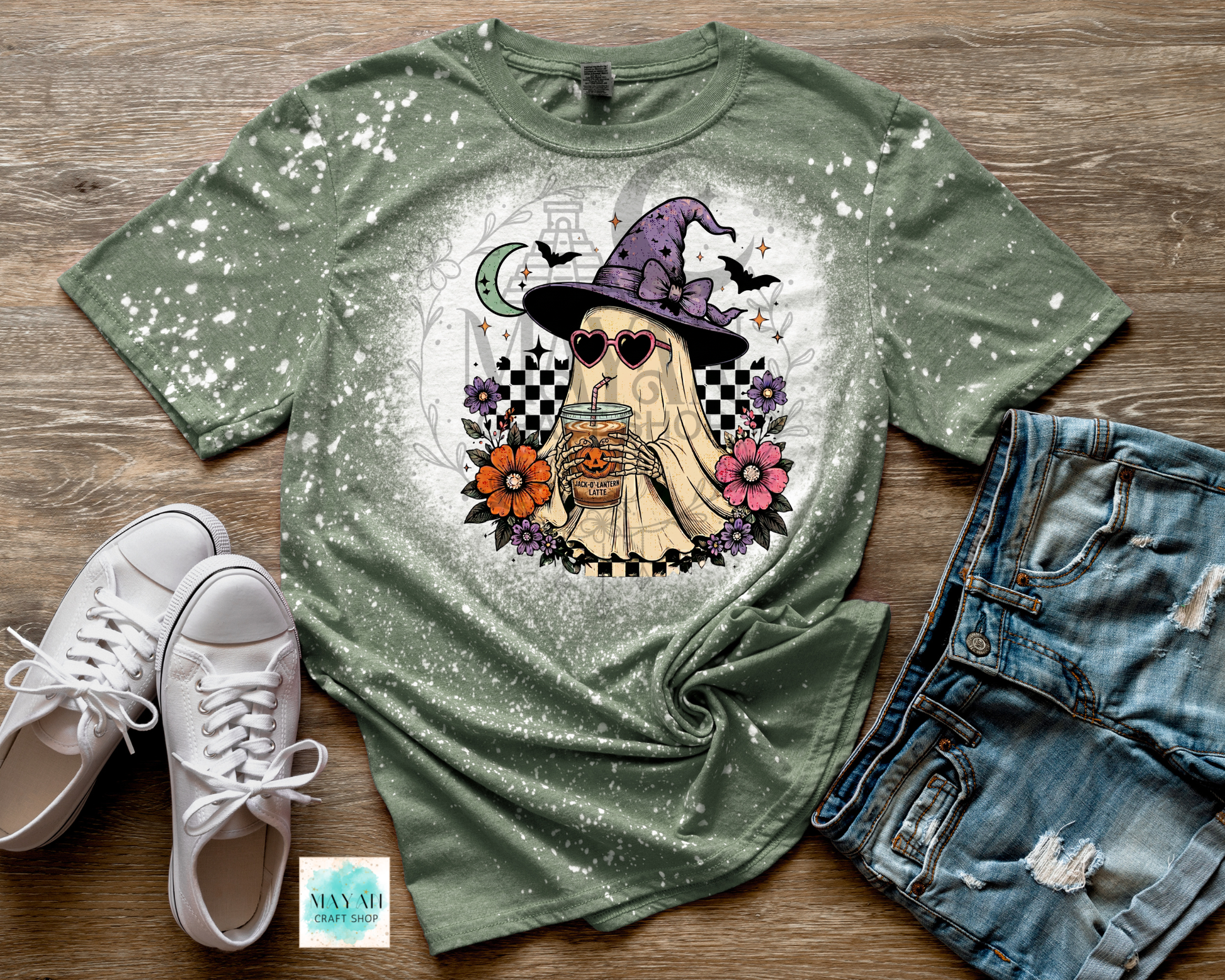 Green t-shirt with a ghost in a witch hat design, paired with white sneakers and denim shorts on a wooden surface.