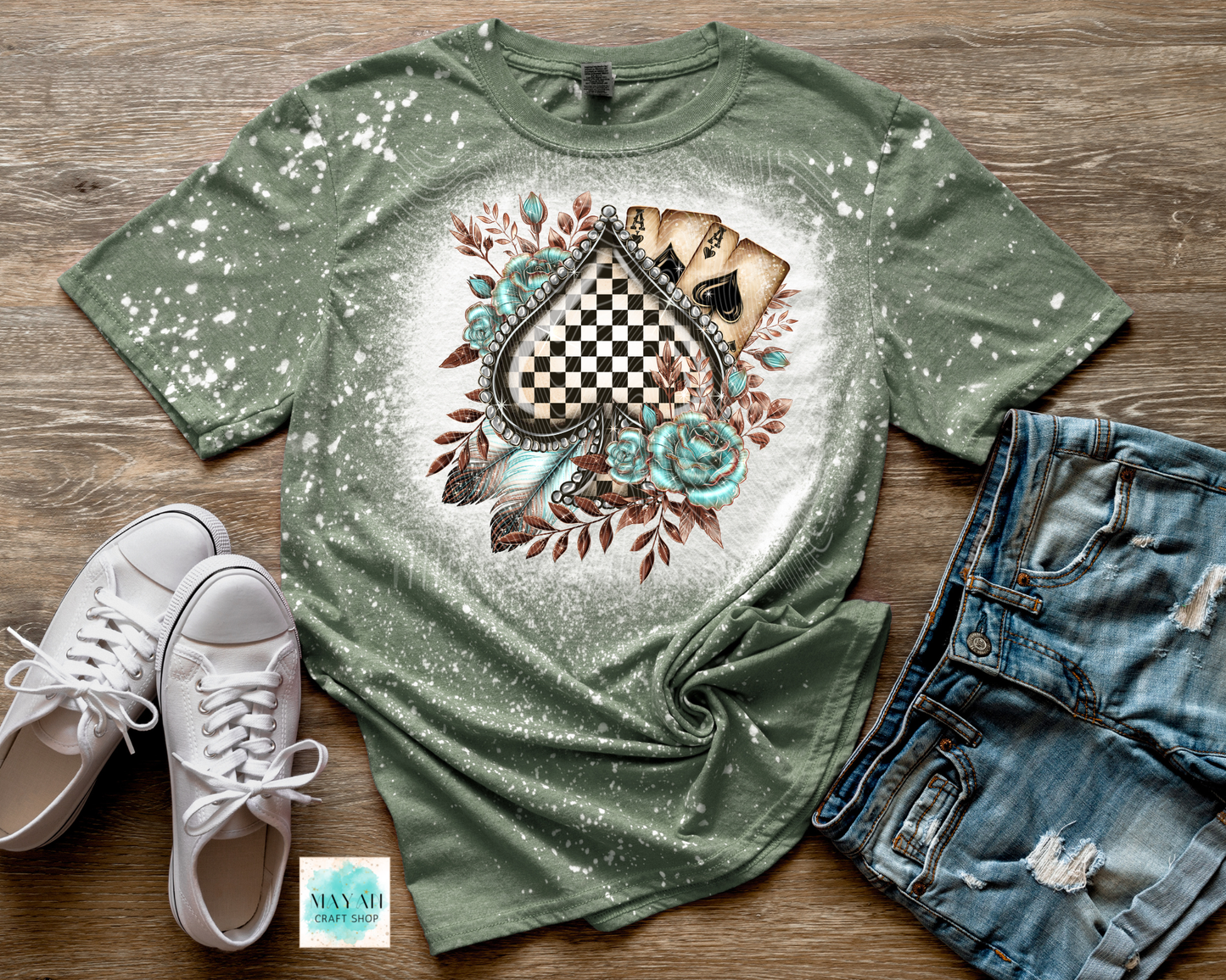 Green t-shirt with a decorative design on a wooden surface with white sneakers and denim shorts.