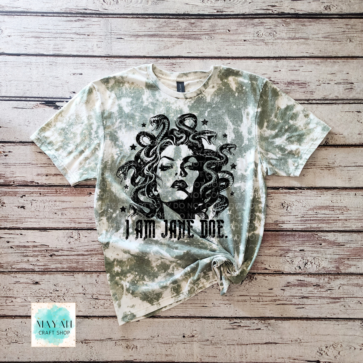 A short-sleeve heather military green bleached graphic t-shirt featuring a black illustration of Medusa and the text 'I AM JANE DOE.' printed on the front, laid flat on a rustic wooden plank background.