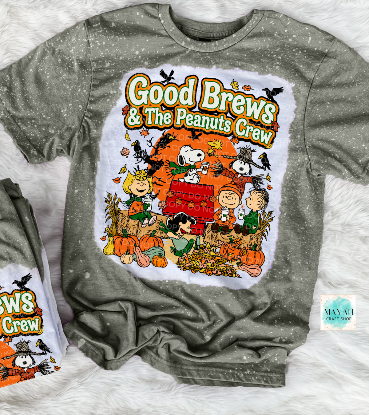 A heather military green bleached t-shirt featuring a large graphic print of the 'Good Brews' with characters in a fall harvest scene. The shirt is laid flat on a white faux fur background next to a folded matching t-shirt.