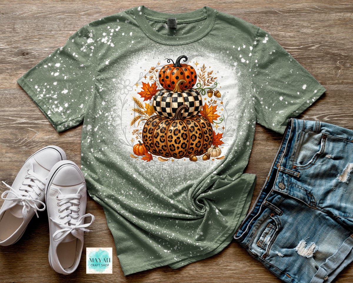 A heather military green short-sleeve t-shirt with a white splatter finish and a central autumn pumpkin graphic, styled in a flat lay with white sneakers, distressed denim shorts.