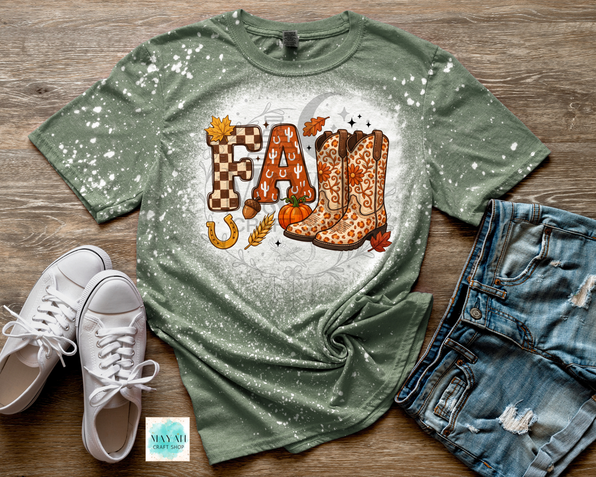 A heather military green short-sleeve t-shirt with a white speckled bleach-dye effect, featuring a 'Fall' graphic with orange patterned cowboy boots, autumn leaves, and pumpkins. The shirt is styled in a flat-lay arrangement on a wooden background alongside white sneakers, distressed denim shorts.